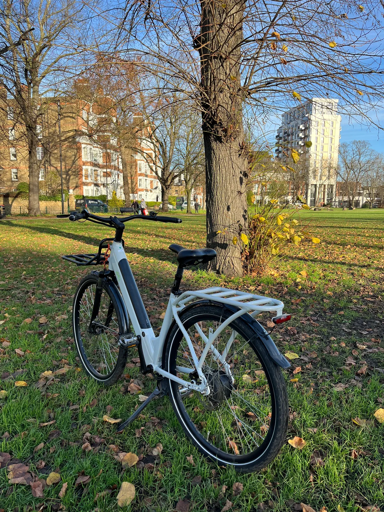 CYCL e-bike parked under a tree in a London park, mansion blocks and a modern tower behind
